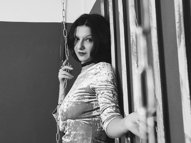 AmethystCharm with short dark hair, wearing a velvet dress, holding a chain, standing against metal bars, looking at the camera with a slight smile, tits visible. Black-and-white photo.