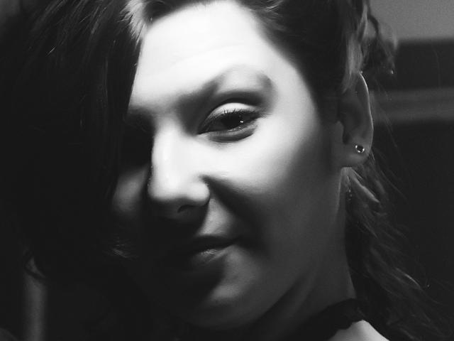 Black-and-white photo of AmethystCharm, showing a partial face with long dark hair covering one eye, smiling slightly, wearing small hoop earrings, and a dark choker.