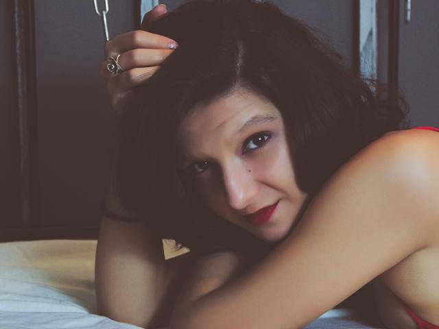 AmethystCharm with dark curly hair, red lipstick, and a ring, laying on a bed, looking at the camera with a slight smile, her arm over her bare tits.