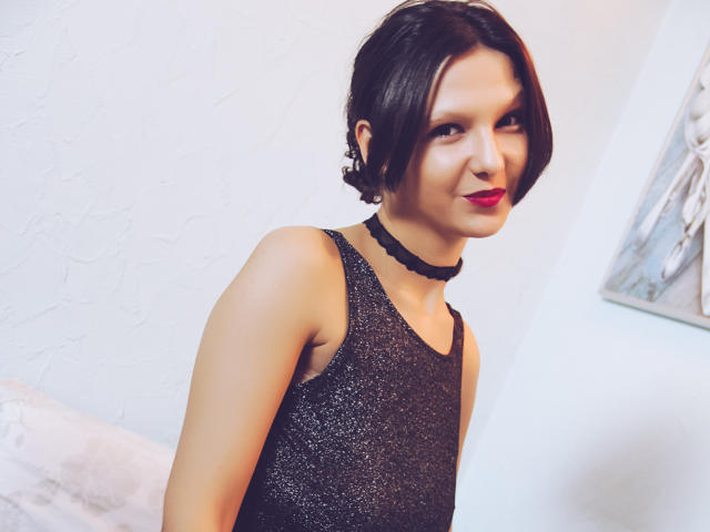 AmethystCharm with short black hair, red lipstick, wearing a sparkly black sleeveless dress and black choker, smiling slightly. Boobs visible, upper body shot.