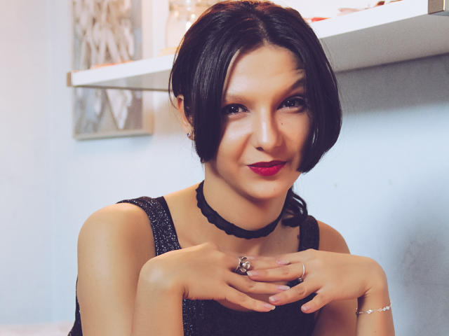 AmethystCharm with dark hair, red lipstick, black choker, and black dress, hands near chest, smiling, wearing ring and bracelet, in a softly lit room.