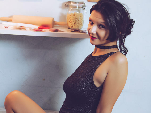 AmethystCharm with dark hair in a messy bun, wearing a sparkly black sleeveless top and black choker, sitting with her ass up, looking at the camera.