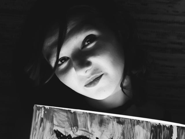 Black-and-white photo of AmethystCharm with soft light on her face, looking up with dark, wavy hair, holding a brush, showing subtle makeup and gentle expression.