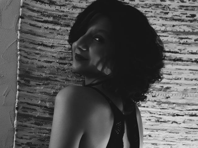 Black-and-white photo of AmethystCharm with short, wavy dark hair, looking over her shoulder, wearing a sleeveless black top, showing her shoulder and part of her face.