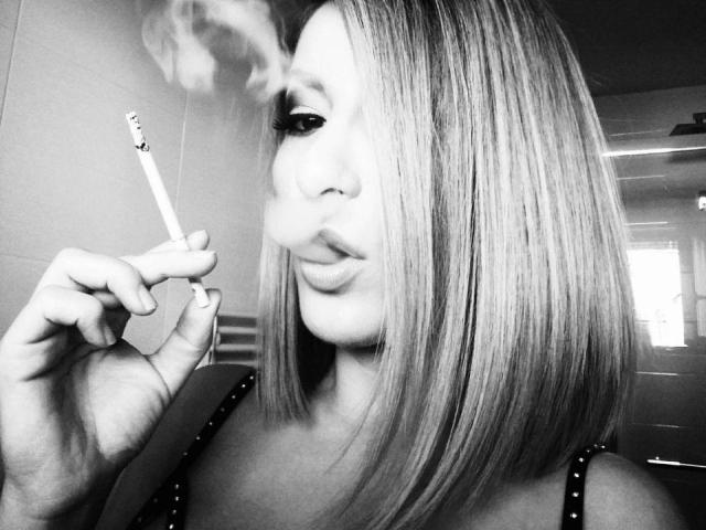 Black and white photo of MayaEva with a bob haircut, smoking a cigarette, her nose pierced, pouting with full lips, wearing a studded top, visible window in the background.