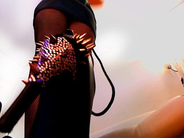 AlfaFemale in a black skirt and fishnet stockings, wearing spiked high heels, raising her leg, showing her shiny brown nails, and a black strap, viewed from a low angle.
