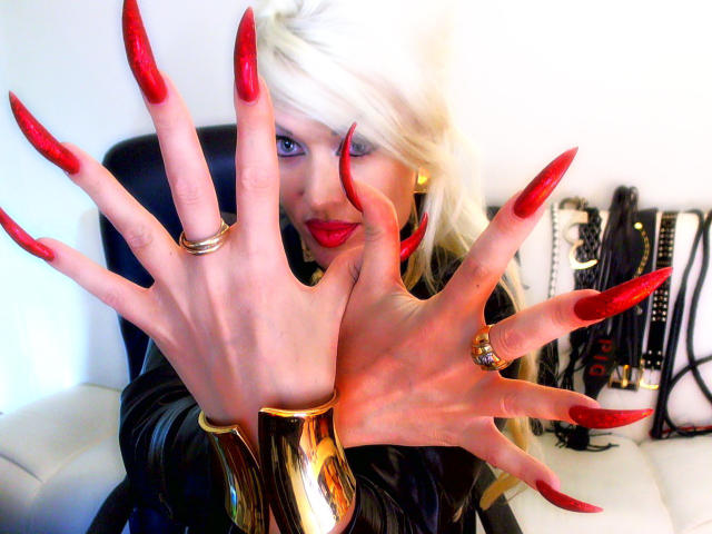 AlfaFemale with long, sharp, red acrylic nails, wearing a black latex outfit and gold rings, reaching forward, showing off her fingers, blonde hair, red lipstick, and a confident smile.