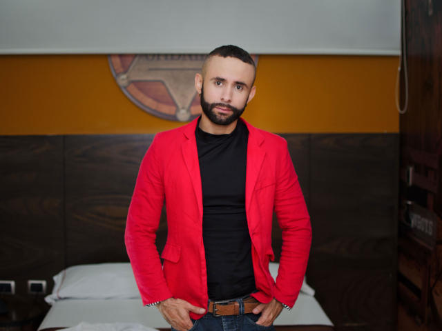 Hij, met een rood jack, een zwart shirt en blauwe jeans, staat in een kamer, er serieus uitzien, met zijn handen op zijn riem, kort donker haar, een baard en een gespierd lichaam..