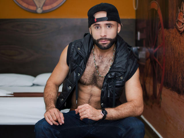 Hevan, sem camisa, usando um colete preto, com um boné virado para trás e um colar, sentado em uma cama, mostrando um peito peludo e braços musculosos, com um relógio no pulso..