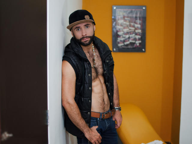 Hevan, zonder shirt, met een zwart gewatteerd vest, blauwe jeans en een zwarte pet, leunt tegen een muur en toont zijn behaarde borst en een serieuze uitdrukking. Hij draagt een ketting..