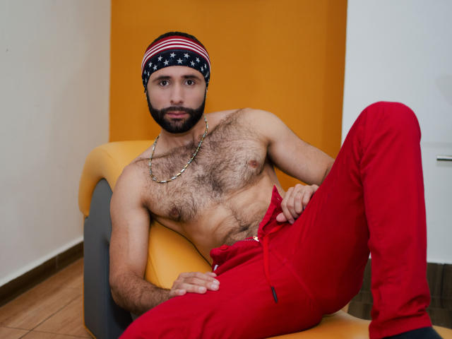 Hevan sem camisa, com peito peludo, calças vermelhas e um lenço com a bandeira americana, sentado em uma cadeira amarela, usando um colar de corrente, olhando para a câmera..