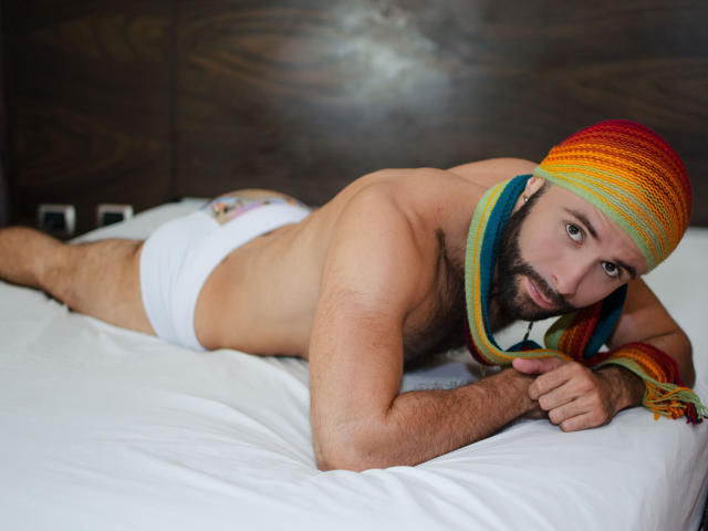 Hevan, met een behaarde borst en baard, ligt op een bed, gekleed in witte onderbroek, een regenbooghoofdband en een bijpassende sjaal, en kijkt met een serieuze uitdrukking naar de camera..