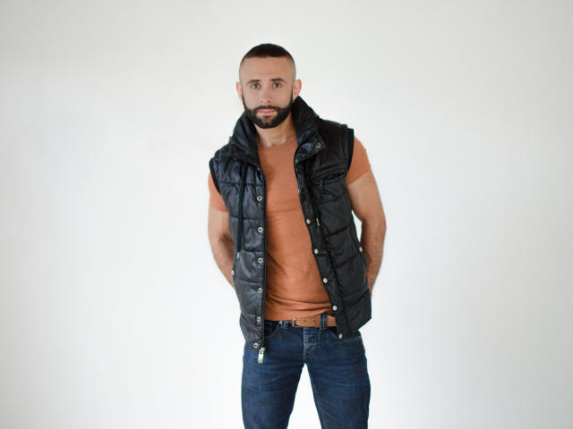 Een gespierde man met baard, zwarte, opvallende vest, oranje T-shirt, blauwe jeans, handen in de zakken, serieuze uitdrukking, kort bruin haar, witte achtergrond, staand, casual outfit..