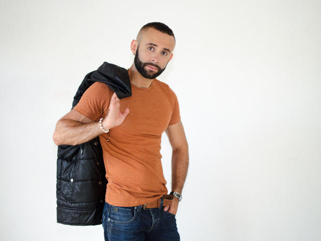 Hevan, met een zwart, gewatteerd vest over zijn schouder, heeft een verraste uitdrukking, draagt een bruin T-shirt, blauwe jeans en een armbandje om zijn rechterpols, en staat tegen een witte muur..