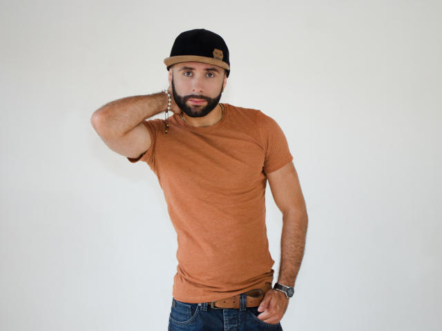 Hevan tem uma camiseta marrom, calças jeans azuis, um boné preto e uma barba, com a mão perto da cabeça, usando uma pulseira de contas e um cinto marrom..