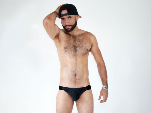 Hevan, sem camisa, usando uma sunga preta, boné preto virado para trás, peito peludo, relógio de pulso, braço direito levantado, mão dentro do boné, olhando para a câmera, físico médio, barba rala, fundo branco..