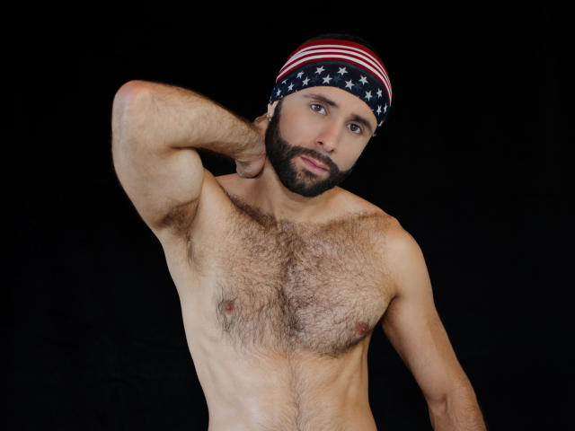 Hevan, com o peito peludo, usando um bandolim com a bandeira americana, tem barba, um braço levantado e uma expressão séria, contra um fundo preto. Ele está sem camisa, olhando para a câmera..