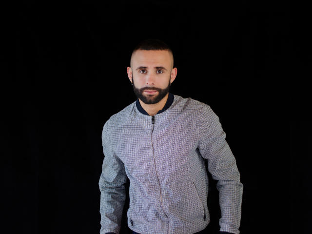 Hevan, com barba aparada, cabelo curto e escuro, vestindo uma camisa cinza xadrez com zíper, em pé contra um fundo preto, com uma expressão de surpresa, olhos arregalados e boca ligeiramente aberta..
