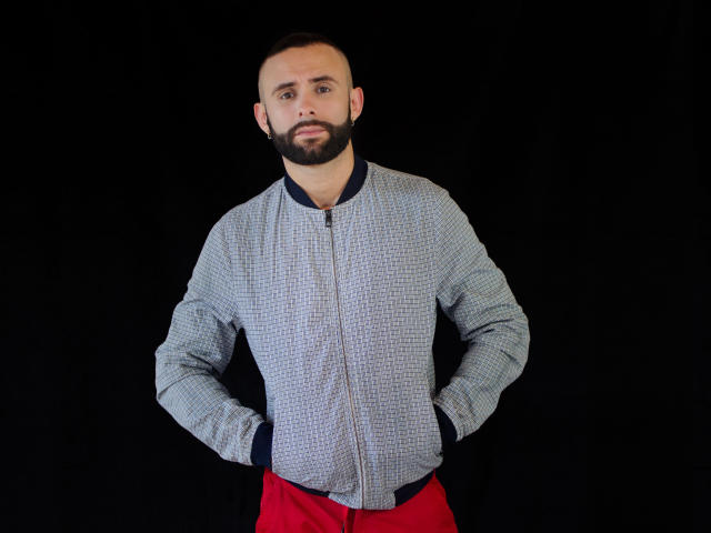 Hevan, een man met een baard en een kaalgeschoren hoofd, draagt een geruit vest met rits en rode broek. Hij staat tegen een zwarte achtergrond, met zijn handen in zijn zakken, en heeft een serieuze blik..