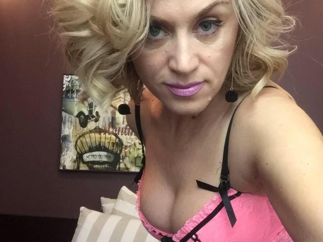 BiankaRay mit blonden Locken, rosa Dessous, schwarzen Trägern, lila Lippenstift und Ohrringen, die ihre Brüste in einem Selfie zeigt und direkt in die Kamera schaut..