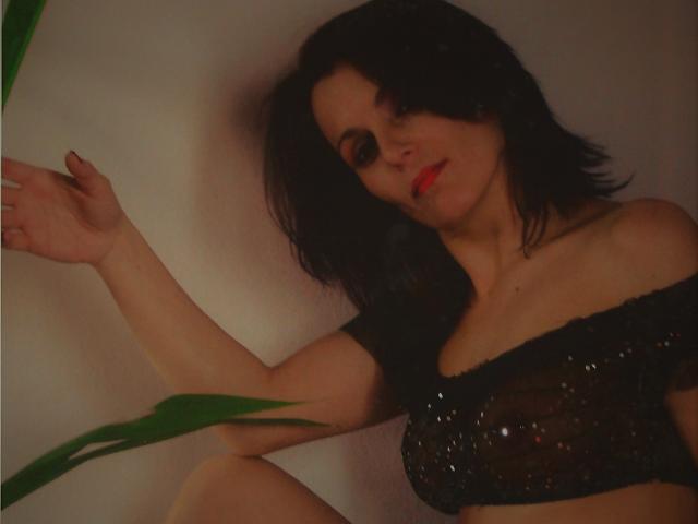MelindaHottX with black hair, red lipstick, and sequined black top, showing her tits, arm raised, green plant in foreground, soft lighting, slightly blurred.