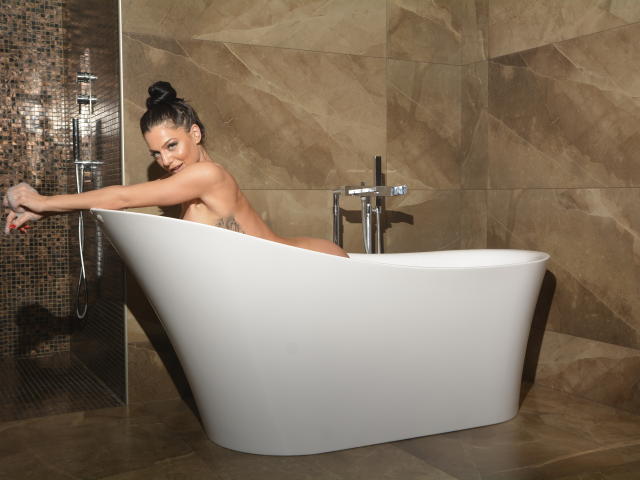 CrystalSel, con cabello oscuro recogido en un moño, desnuda, en una bañera blanca, estirándose con los brazos extendidos, mostrando sus pechos y glúteos, baño con paredes de mármol marrón, mirando a la cámara..
