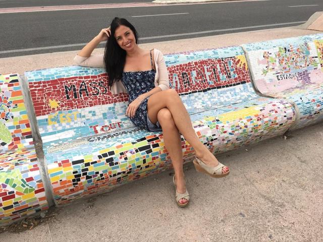 KatieFrenchie, with long black hair, sits on a colorful mosaic bench, wearing a blue floral dress, beige cardigan, and tan wedge sandals, smiling with legs crossed.