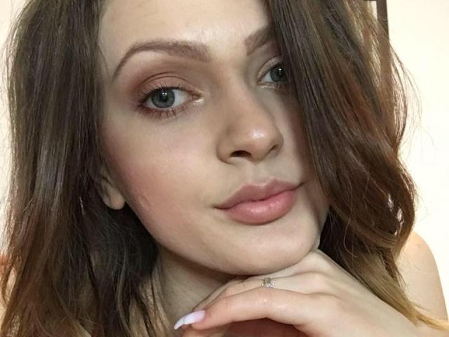 Le visage de BlossomPussy, avec ses cheveux bruns ondulés, son maquillage nude rosé, ses yeux bleus, ses lèvres roses, ses joues légèrement rougies, le menton reposant sur sa main, portant une simple bague, a l'air adorable..