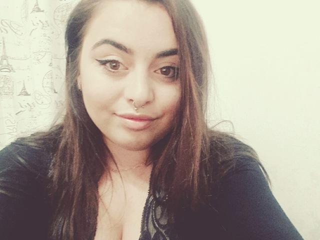 ChaudePourxToi has brown eyes, a nose ring, and winged eyeliner, with long brown hair and a slight smile, wearing a black top, showing a hint of cleavage.