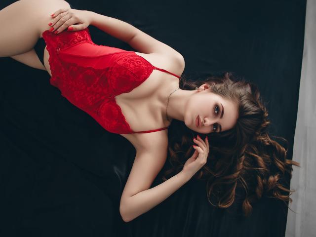 ShyNaomi è sdraiata su una superficie nera, indossando una body in pizzo rosso che mette in mostra il seno e i glutei. Ha lunghi capelli castani, unghie rosse e un'espressione seria..