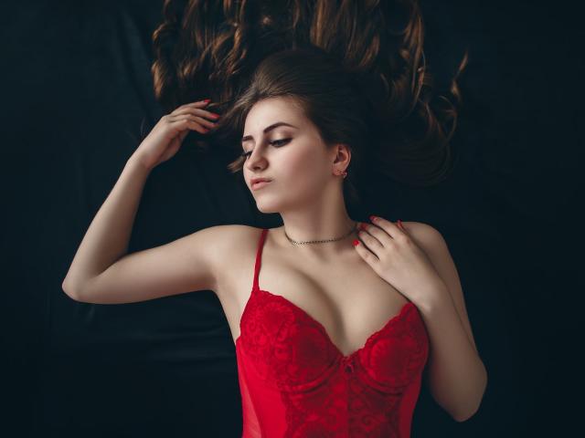 Schüchterne Naomi mit langen braunen Haaren, roter Spitzendessous, und einer Halskette, liegt, Augen geschlossen, eine Hand auf der Brust, die andere Hand im Haar, blasse Haut, rote Nägel, dunkler Hintergrund..