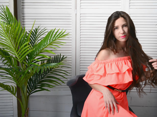 RoyalBonie with long brown hair, wearing an off-shoulder neon pink dress, sitting on a black chair, next to a green plant, has pink lipstick and a subtle smile.