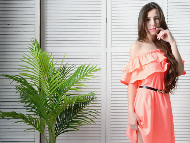 RoyalBonie, con cabello largo y castaño, vestida con un vestido naranja de corte asimétrico, un cinturón negro y uñas rosas, está de pie junto a una planta verde, tocándose suavemente los labios..