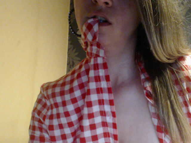 AngryGirl69 mit einem rot-weißen Karoshirt, die sich die Unterlippe anbeißt und dabei ein wenig ihrer Brust zeigt, mit langen blonden Haaren, teilweise im Gesicht zu sehen, und mit offenem Mund..
