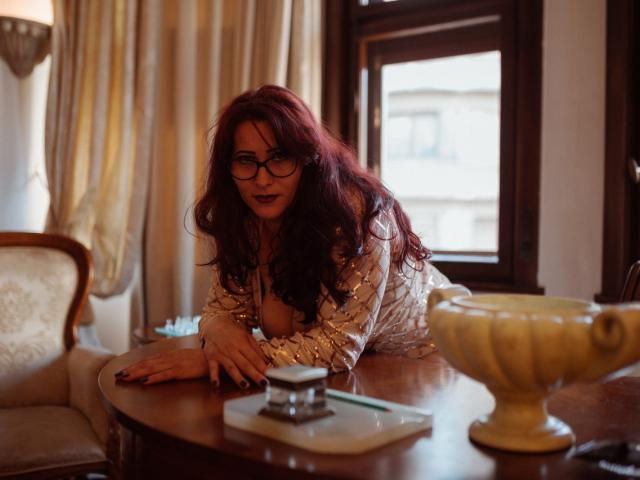 SoniaLaBelle with red hair, glasses, and cleavage, leaning on a wooden table, wearing a sheer, patterned top, in a warmly lit room, with a yellow vase in front.