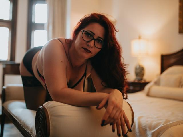 SoniaLaBelle with red hair, glasses, and black lingerie, leaning on a bed, showing her tits and ass, in a warmly lit bedroom, looking at the camera.