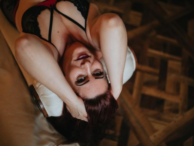 SoniaLaBelle with red hair, black lace bra, arms raised, hands behind head, looking up, showing her tits, lying on white chair, wooden floor, purple lipstick, smoky eyes.