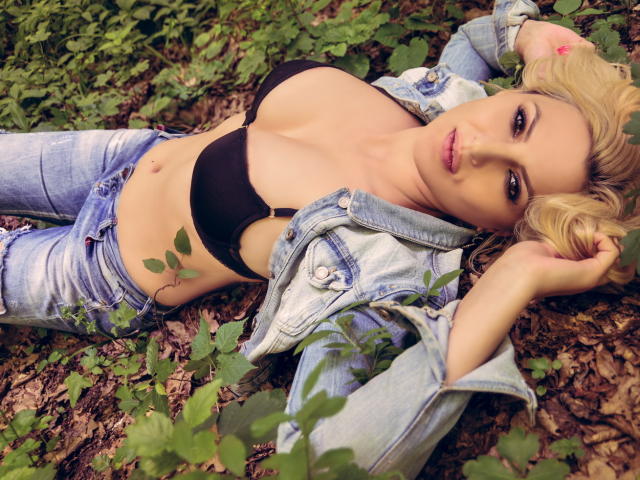 SaraLongLegs laying in the dirt, wearing a black bra that shows her big boobs, a denim jacket, and faded jeans, with green leaves around her. Blonde, sexy, and relaxed.