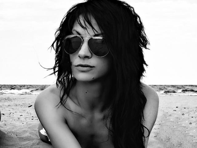Black-and-white photo of CassandraMichelli with wet, dark hair, wearing aviator sunglasses, and nothing on, sitting on a sandy beach, looking serious with a slight pout.