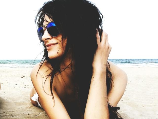 CassandraMichelli with wet black hair, wearing blue aviator sunglasses, smiling, hands in hair, topless, sandy beach, ocean in background, sexy, relaxed, natural light, casual vibe.