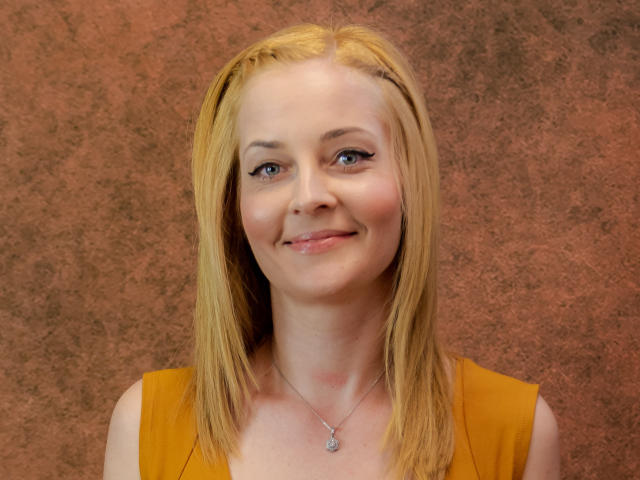 SoleildeNuit has blonde hair, blue eyes, and a slight smile. She's wearing a yellow top and a silver necklace with a small pendant. Her tits are small and cute.