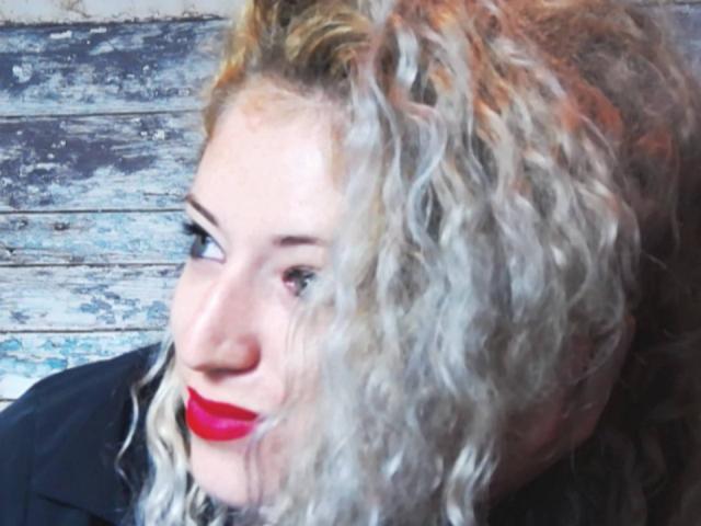 SassyKate with curly blonde hair, red lipstick, and a black top, looking sideways with a slight smile, showing her nose and part of her eye.