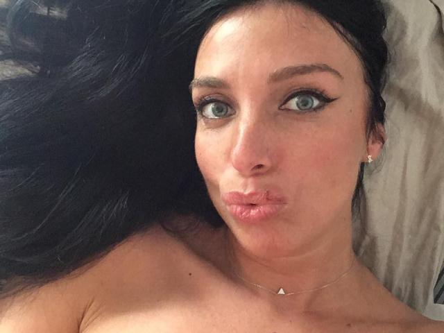 KatieFrenchie with pouty pink lips, blue eyes, and black hair, wearing a gold necklace, lying down, looking at camera, her bare shoulders visible.