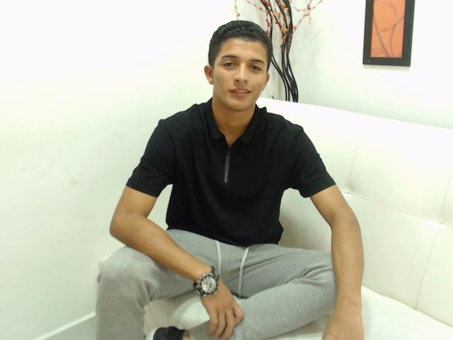 NicolasSexy, een jonge Latino man met kort zwart haar, gekleed in een zwarte hoodie en grijze broek, zittend op een witte bank, lachend, met een horloge om zijn linkerhand..