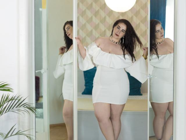 LexyRose with large tits, pale skin, and dark hair, wearing a white off-shoulder dress, stands in front of a mirror, her reflection showing her curvy ass.