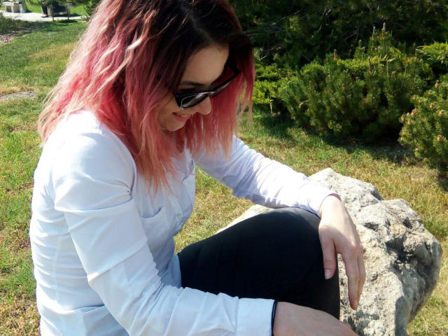 AliciaKristal mit rosa gefärbtem Haar, schwarzen Sonnenbrillen, einem weißen Hemd und einer schwarzen Hose, sitzt auf einem Felsen, berührt ihn mit ihrer rechten Hand und blickt nach unten..