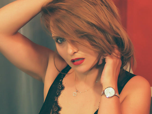 Oliviaaa, avec du rouge à lèvres, un haut en dentelle noire et un fond rouge, a une main dans ses cheveux bruns en désordre, porte une montre et un collier en argent, et a une allure séduisante..