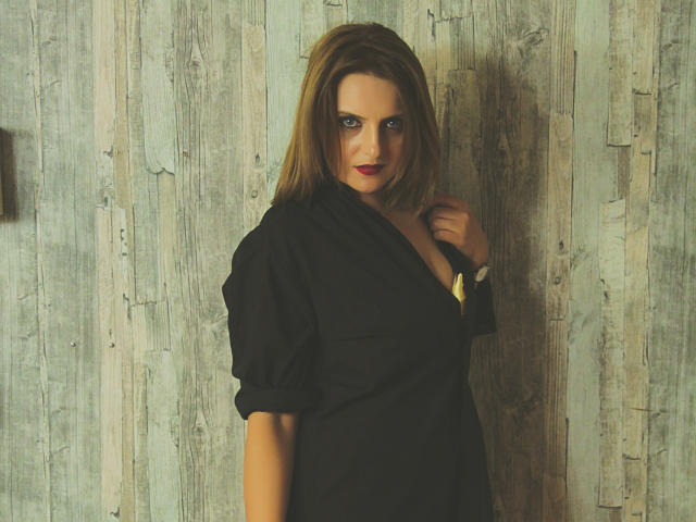 Oliviaaa, avec une poitrine de taille moyenne, portant une chemise noire, debout devant un mur en bois, une main près du cou, rouge à lèvres, yeux bleus et cheveux blonds. Son expression est sérieuse et confiante..