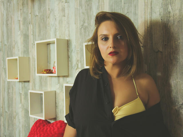 Oliviaaa with shoulder-length brown hair, red lipstick, black shirt partially open, yellow bra showing small tits, standing against wooden wall, white shelves in background.