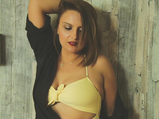 Oliviaaa with red lips, yellow bikini top showing her tits, black shirt open, brown hair, arm raised, wood background, subtle necklace, sexy, confident, slight smile.